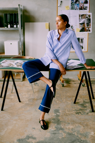 Woman sitting on a table in a room with shelves and posters. b. young BYHanio Tape Shirt