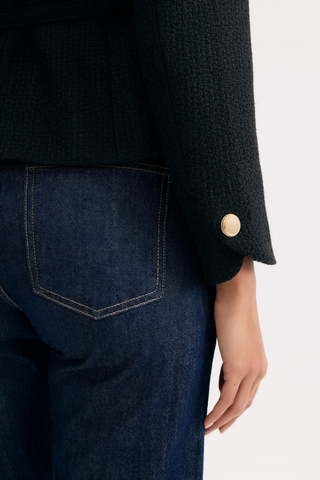 Leo & Ugo KE376 Salomé Elegant Belted Jacket - Black  Close-up of a person wearing a black coat with a gold button and dark blue jeans on a white background.