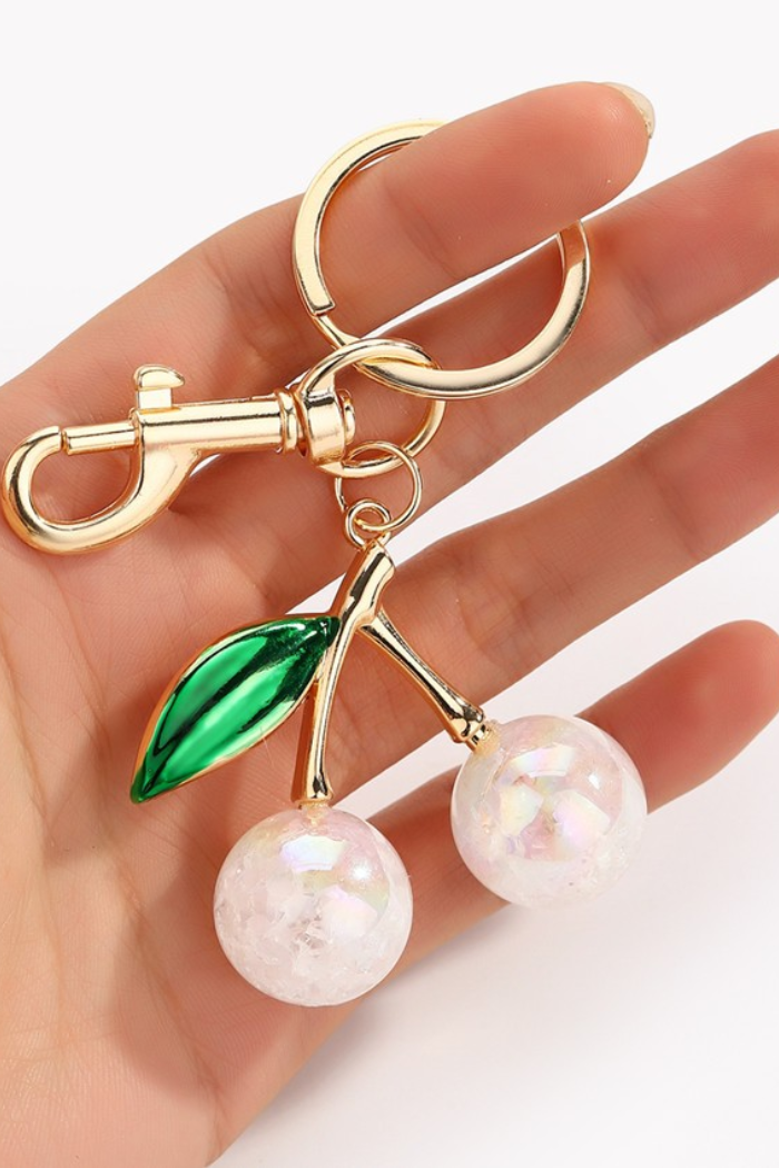 Close-up of a sparkling cherry keychain featuring two clear, iridescent glitter-filled cherries, a shiny green enamel leaf, and gold-tone metal hardware (keyring and lobster clasp) on a light blue background.