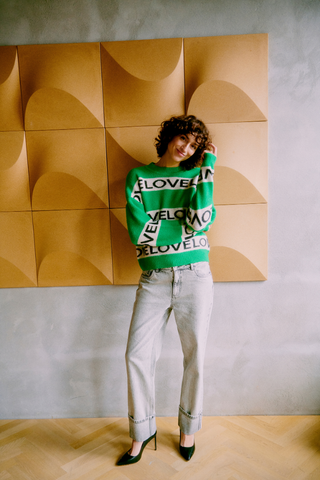 Person wearing a green sweater with text, standing against a wall with geometric brown panels. b. young BYOmea Love Sweater