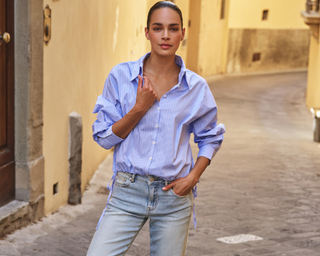 Person wearing a light blue shirt and jeans standing on a stone street.