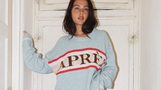A woman wearing an oversized light blue knit sweater with 'APRÈS' in brown and red stripes, paired with textured brown wide-leg pants, standing in a rustic hallway.