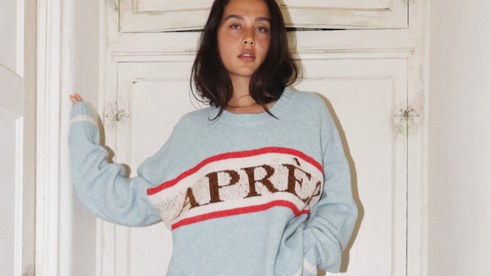 A woman wearing an oversized light blue knit sweater with 'APRÈS' in brown and red stripes, paired with textured brown wide-leg pants, standing in a rustic hallway.