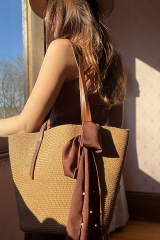 Leo & Ugo KEBH087 Pearl Detail Hat & Bag Combo Woman holding a straw bag with a brown bow, standing against a neutral wall.