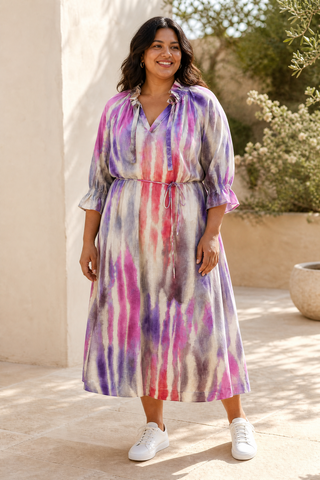 A woman stands outdoors wearing a midi-length tie-dye dress with purple, pink, and white vertical streaks, paired with white sneakers. The background shows a warm beige stucco wall and sunlit garden plants.
