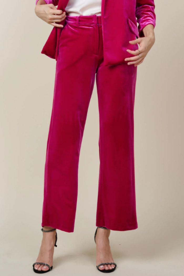 Full-length image of women's fuchsia pink velvet wide-leg trousers, styled with a matching velvet blazer and black high heels with delicate ankle straps, on a light beige background.

