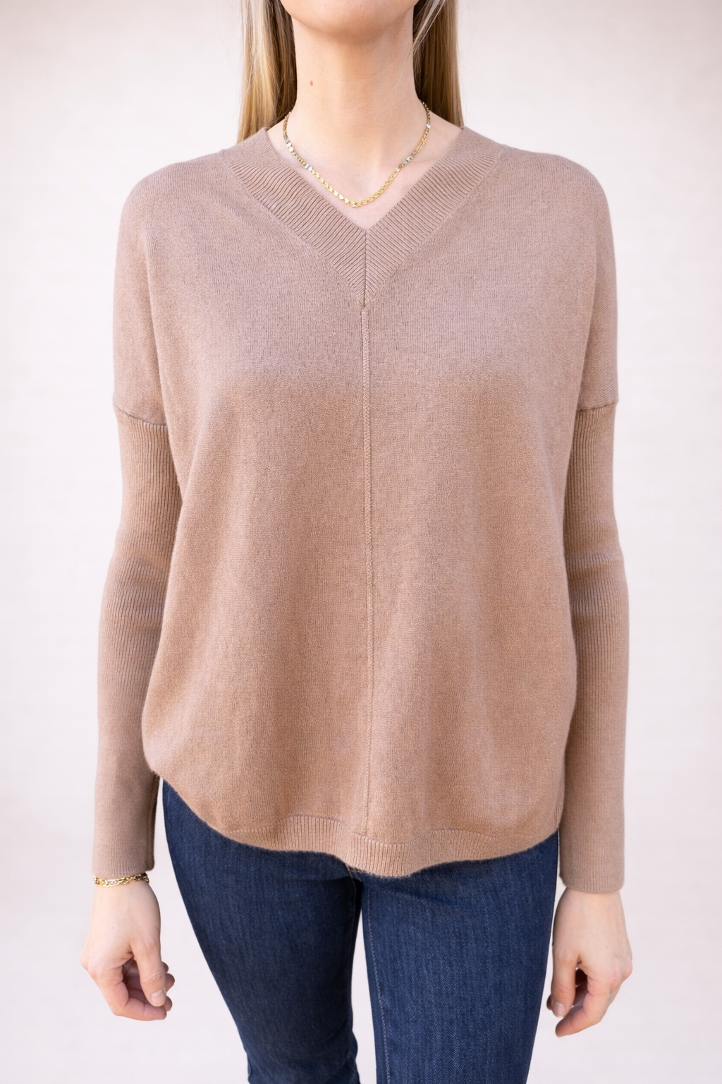 A model wears a light brown (taupe) women's relaxed fit V-neck sweater, made from soft knit fabric, paired with dark blue jeans, standing against a neutral background.
