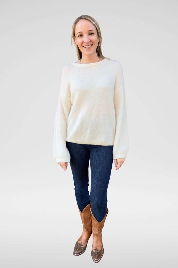 Smiling woman wearing a relaxed fit cream boat neck knit sweater paired with dark jeans and brown cowboy boots on a light background.