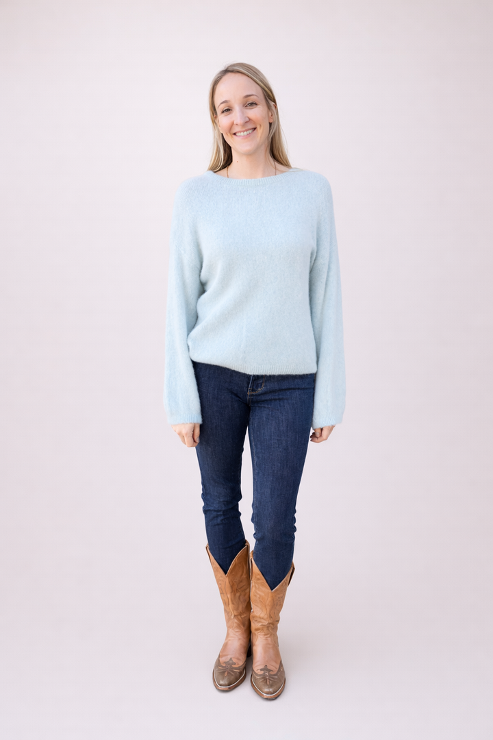Full shot of a smiling woman wearing a light blue fuzzy boat neck sweater with long sleeves, dark blue jeans, and tan cowboy boots, standing against a light background.