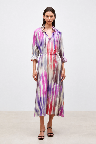 A model stands against a plain light gray studio background wearing a long-sleeve midi dress with a tie-dye pattern in purple, pink, red, and blue on a cream base, paired with black strappy sandals.