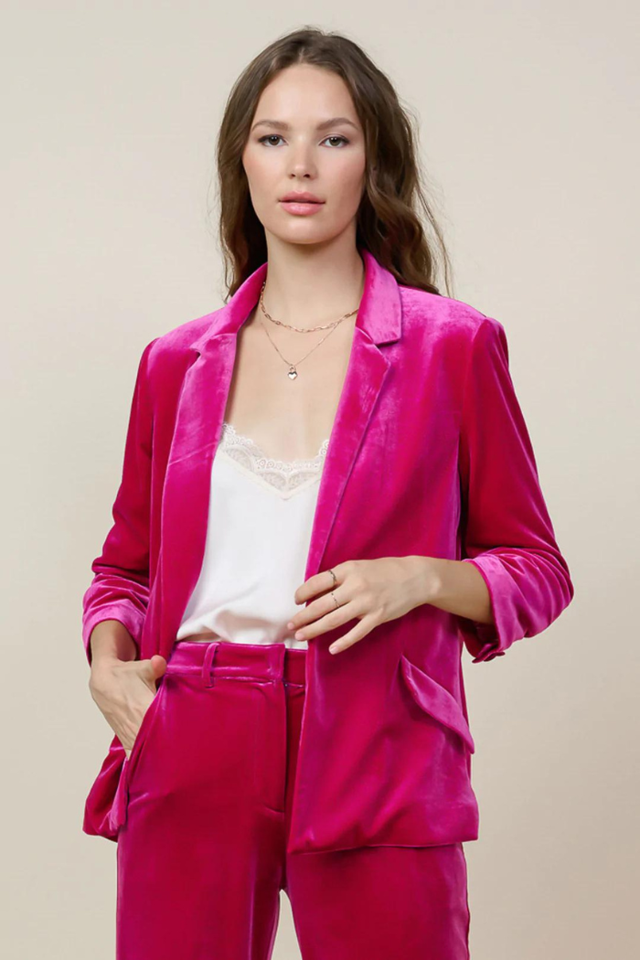 A stylish woman wearing a vibrant fuchsia pink velvet tailored blazer and matching trousers, styled with a white lace camisole and delicate gold necklaces, against a neutral background.