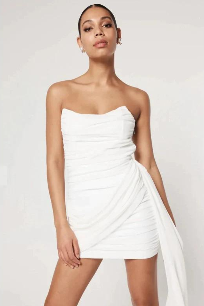 Full-body shot of a woman with dark hair pulled back, wearing an elegant white strapless ruched bodycon mini dress with a sweetheart neckline and a long, flowing draped fabric accent on the right side, against a clean white background.
